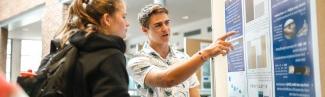 A student points to their presentation poster as they talk to a fellow student